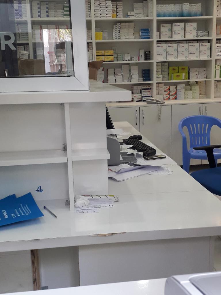 In front of shelves full of boxes of medication is a white desk with a computer keyboard and stacks of paper - a pharmacy.