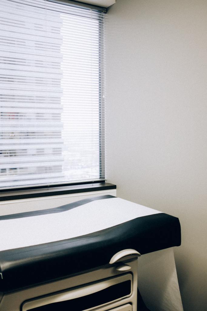 In front of a window with partially closed horizontal blinds there is an examination table/bed with a padded surface covered in navy blue plastic leatherette type material. A roll of white paper has been laid on top of the table.