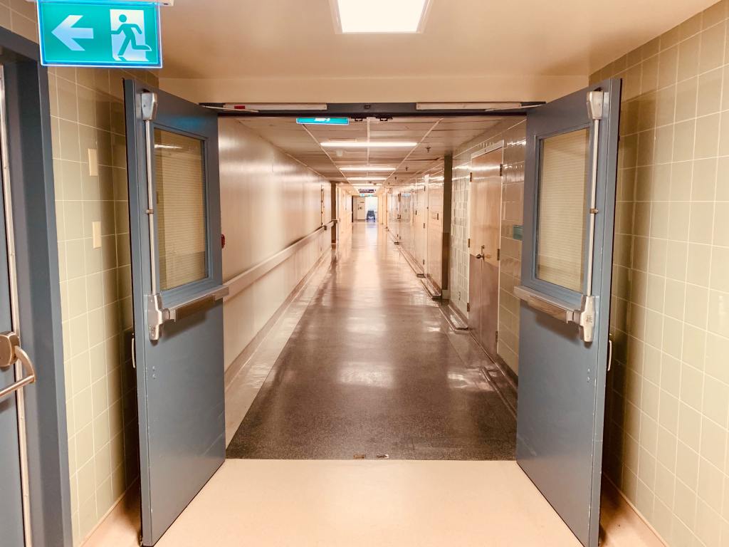A long corridor in shades of grey, white and pale blue. There is artificial lighting which reflects off the polished linoleum floors, and in the foreground fire doors are propped open. There is an emergency exit sign illuminated and pointing to the left.