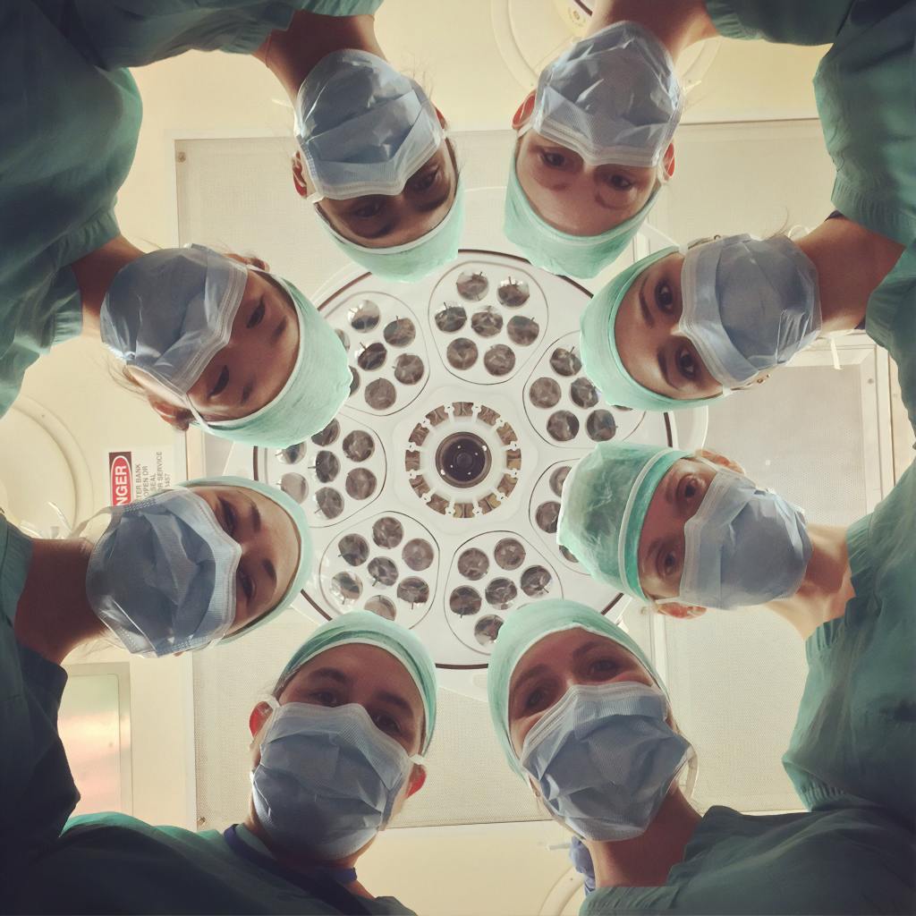 A circle of eight faces in surgical scrubs, hats and masks are arranged in a circle looking down on the camera. Behind them, a round surgical light with many bulbs is visible.
